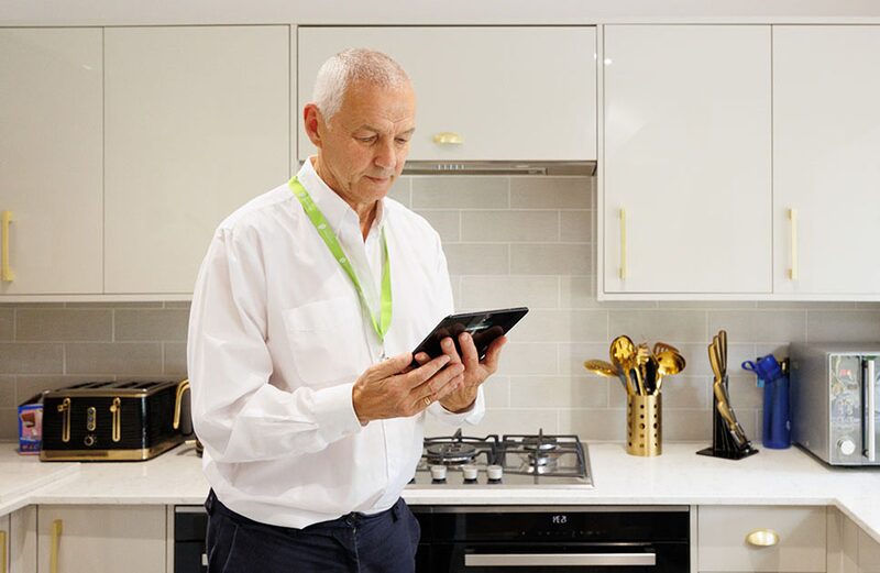 An older man with a green lanyard using a tablet in a modern cream kitchen, apparently an energy assessor carrying out a survey