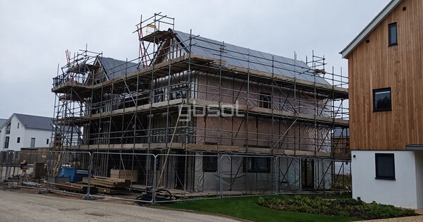 A new-build house under construction wrapped in scaffolding with solar panels being fitted on the roof, next to a finished timber-clad home