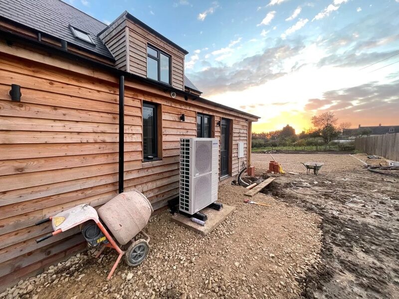 An air source heat pump unit installed on a concrete base outside a timber-clad new-build house at sunset, with a cement mixer and building site in the foreground