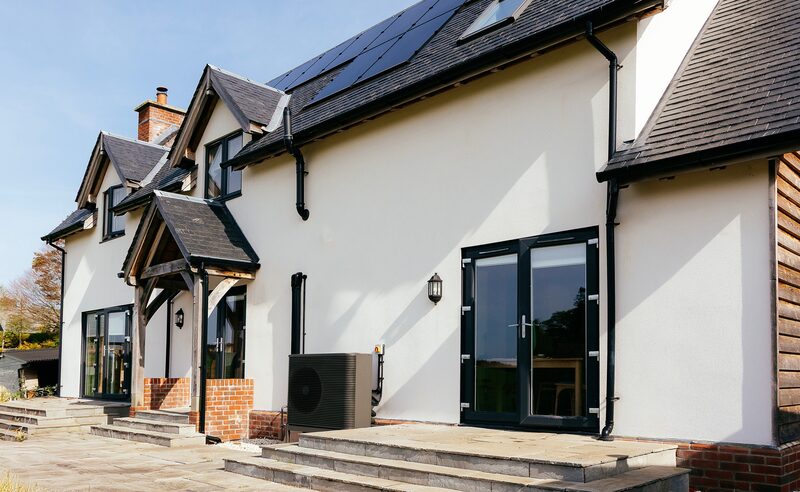 An air source heat pump unit installed on the exterior wall of a modern white-rendered house with solar panels on the roof