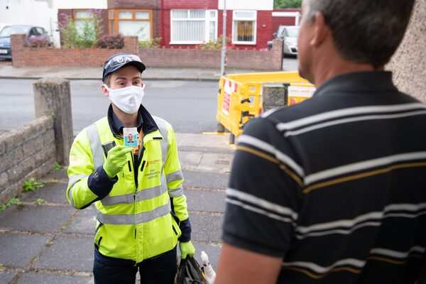 A gas emergency engineer in a high-visibility jacket and face mask shows his ID card to a householder at the door