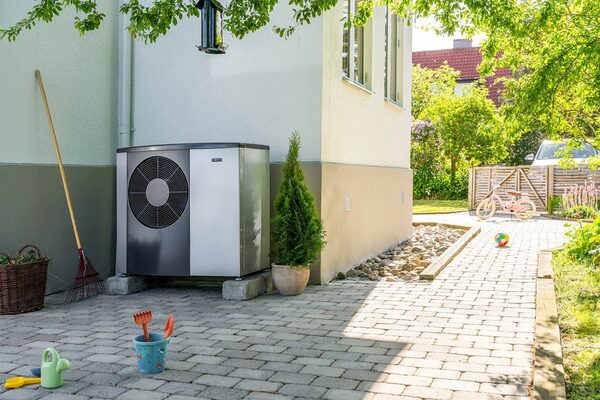 An outdoor air-source heat pump unit installed on a paved driveway beside a house in a sunny garden