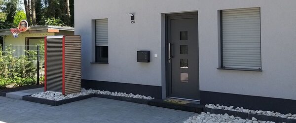 An outdoor air source heat pump unit with a wooden slat cover beside the front door of a modern grey house