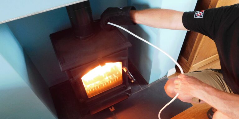 A technician in gloves using a probe cable to check a lit wood-burning stove installed in a fireplace
