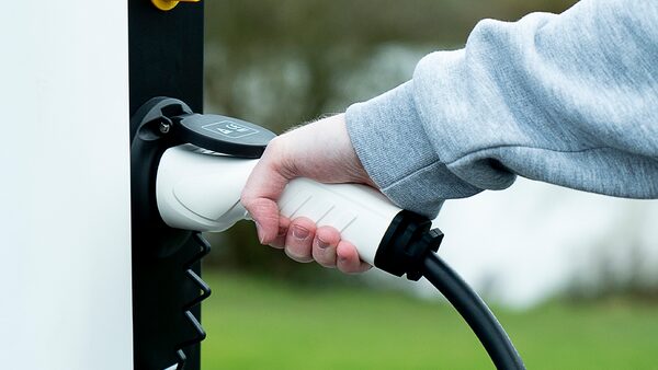 A hand plugging a white charging cable into a wall-mounted electric vehicle charge point outdoors