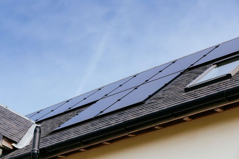 Solar panels fitted flush on the slate-tiled roof of a house with a skylight, seen against a blue sky