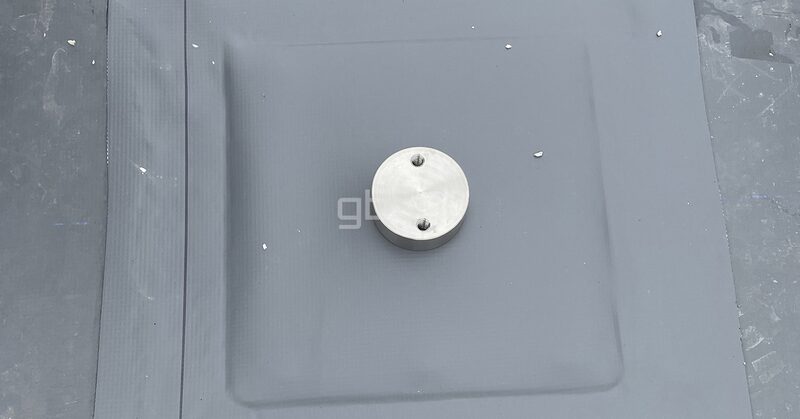 A close-up of a round metal mounting plate fixed to grey flat roofing membrane, part of a solar panel roof mounting detail