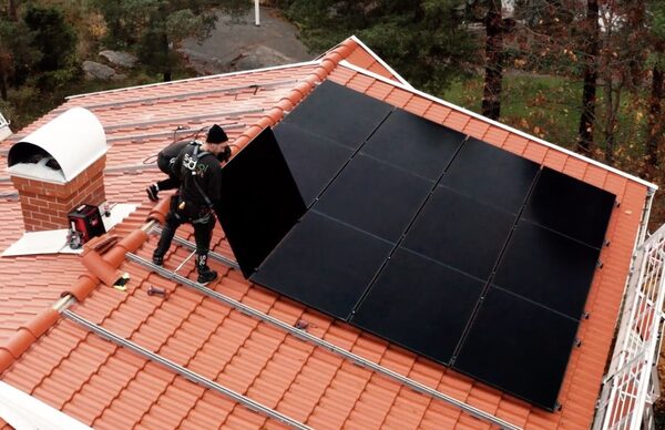 An installer on a terracotta-tiled roof fitting black solar panels alongside an array already mounted