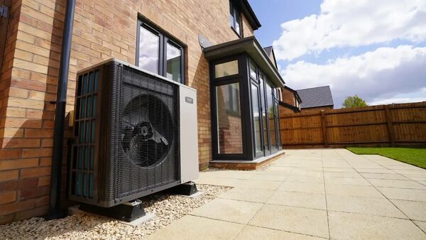 An air source heat pump unit installed on gravel against the brick exterior wall of a modern UK house with a conservatory and paved patio