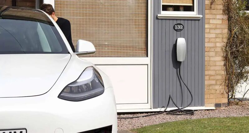 A white electric car parked outside a house with a wall-mounted EV charge point and cable on the grey cladding