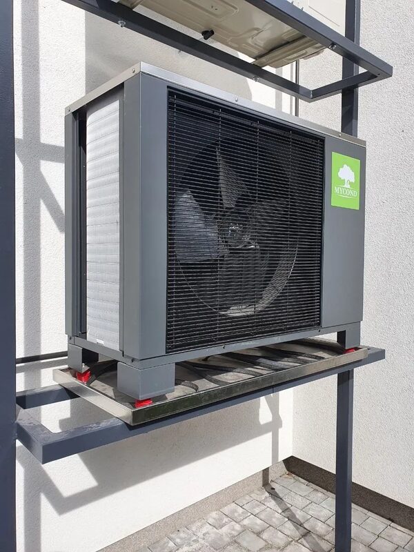 A Mycond air source heat pump outdoor unit mounted on a metal frame against a white house wall
