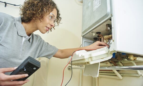 A woman engineer with safety glasses servicing the inside of a wall-mounted gas boiler while holding a multimeter