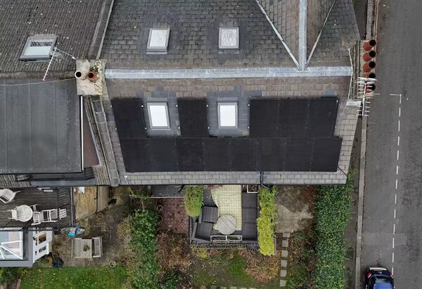 An aerial view of a grey slate roof with black solar panels installed around two skylights, beside a road