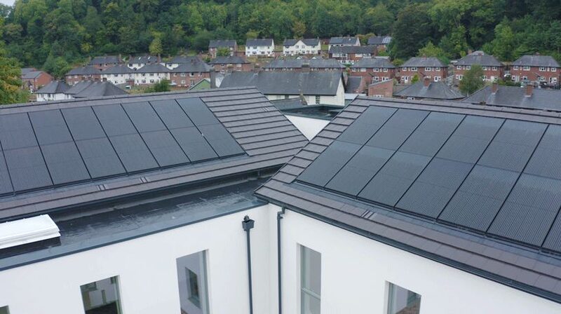 Rooftop solar panels installed on the tiled roofs of a new-build block of flats, with houses and wooded hills behind
