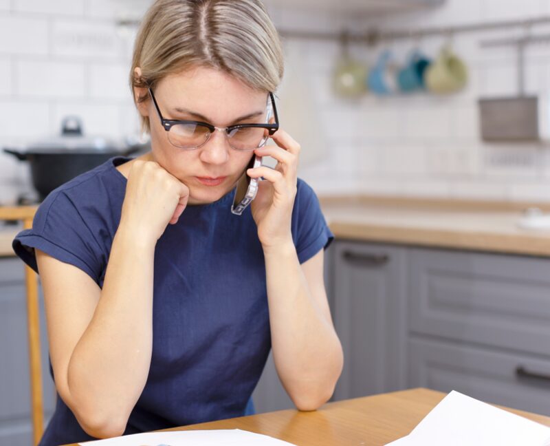 A woman with glasses on the phone at a kitchen table with papers, looking concerned