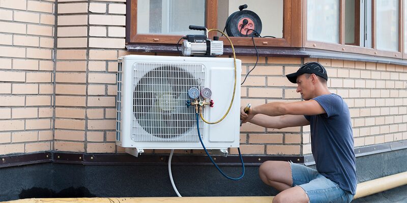 A technician working on an air source heat pump unit mounted on an exterior brick wall of a house