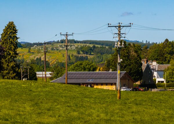 Wooden electricity poles and overhead power lines crossing a green rural field, with a barn roof fitted with solar panels and houses in the background