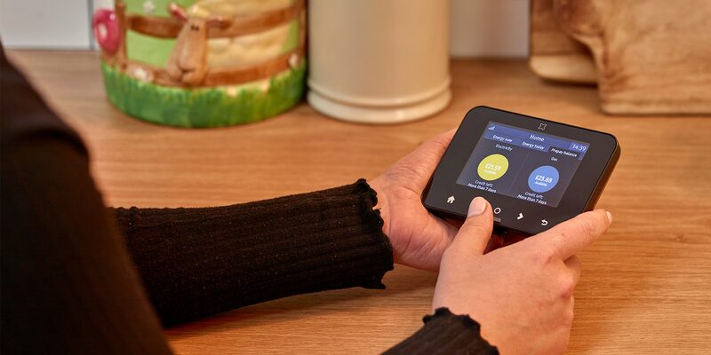 A person holding a smart meter in-home display showing electricity and gas costs on a kitchen worktop