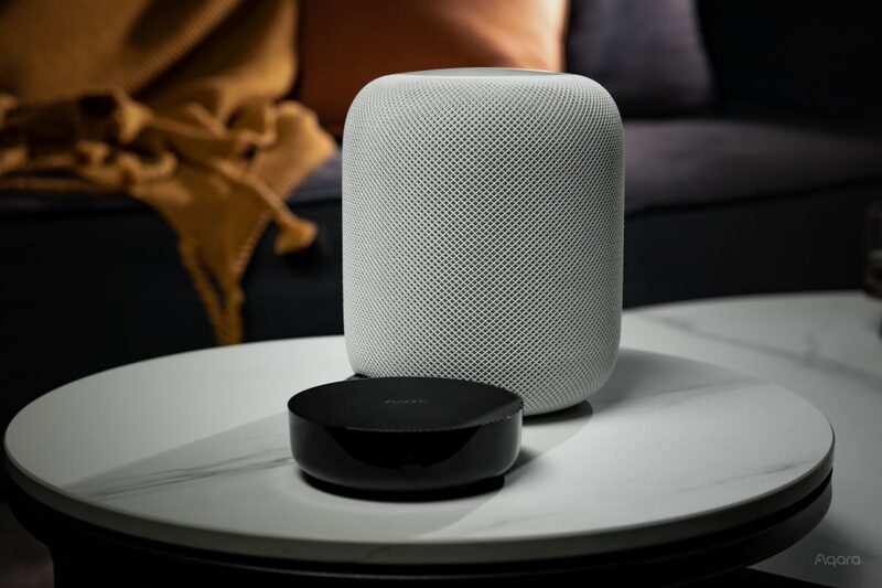 A black Aqara smart hub sitting on a marble table in front of a white Apple HomePod smart speaker in a living room