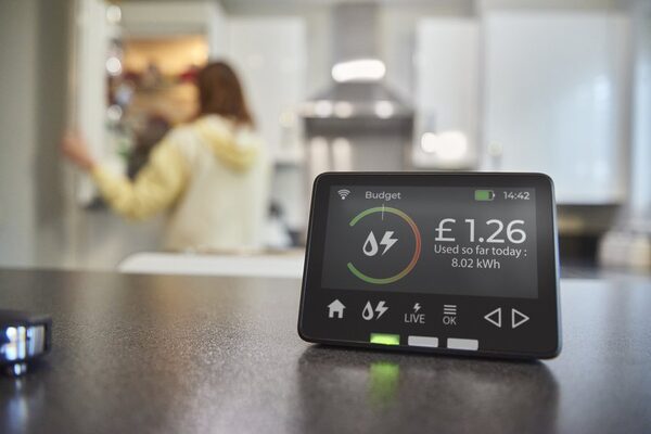 A smart energy monitor on a kitchen worktop showing today's energy spend of £1.26 and 8.02 kWh, with a blurred person in the background