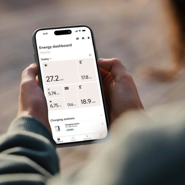 A person holding a smartphone showing the Smappee energy dashboard app with kWh figures and a charging station status