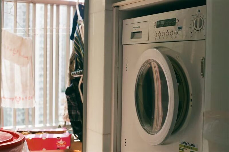 A white front-loading washing machine installed in a small utility space next to a balcony window