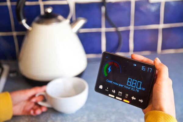 A hand holding a smart energy monitor showing live electricity cost in a kitchen beside a kettle and cup