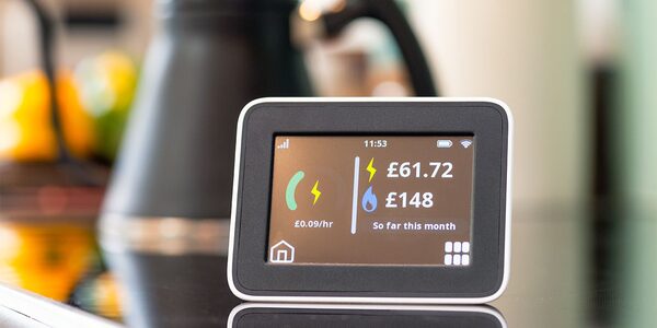 A smart energy monitor in a kitchen displaying electricity and water charges of £61.72 and £148 so far this month at £0.09/hr