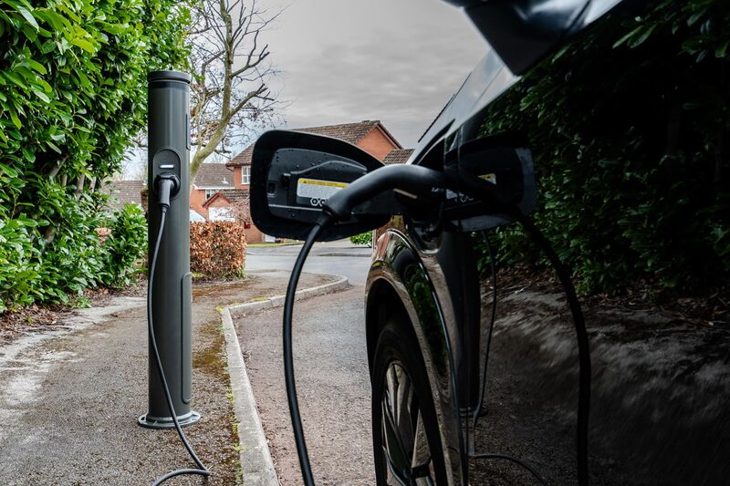 A dark grey pillar-style EV charge point on a residential driveway with a cable plugged into a black electric car