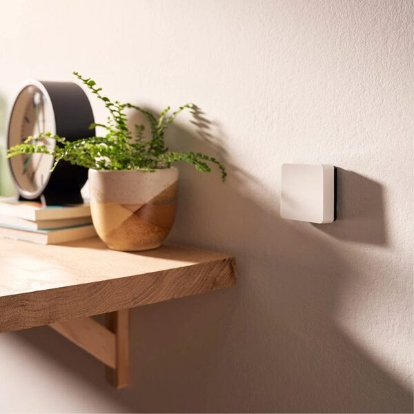 A small white square smart thermostat mounted on a wall beside a wooden shelf with a plant, clock and books