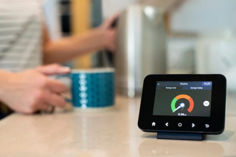 A smart meter display showing energy usage on a kitchen worktop with a person making a drink in the background