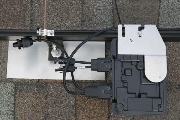 An Enphase microinverter mounted on solar panel racking over asphalt roof shingles, with cabling and connectors