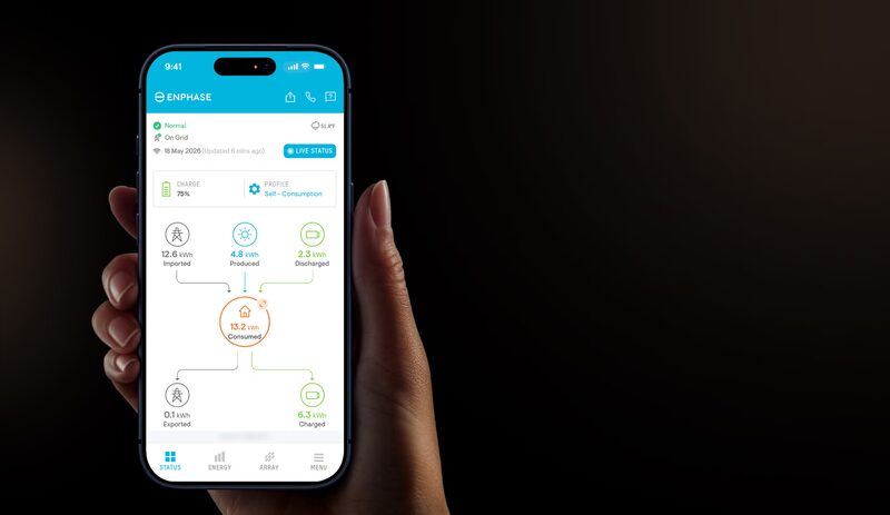 A hand holding a smartphone displaying the Enphase app home energy monitoring screen with solar production, consumption, battery and grid flow figures