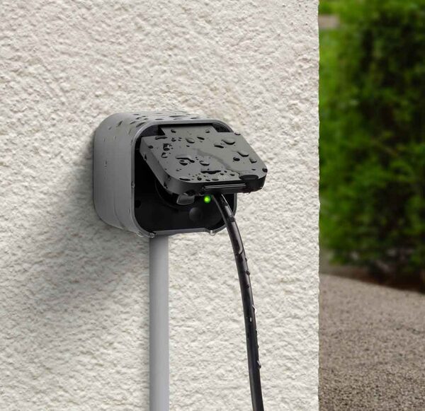 An Eve Energy Outdoor smart plug with a cable plugged in, mounted on an exterior wall with water droplets on it