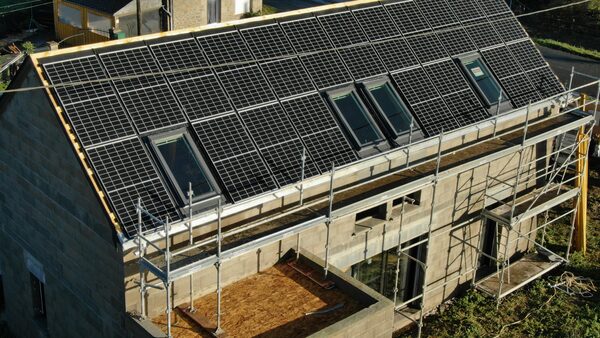 A house under construction with a large in-roof solar panel array covering the roof, surrounded by scaffolding