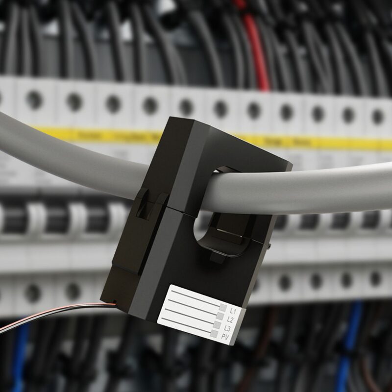 A black CT current clamp fitted around a grey cable in a consumer unit, with L1 L2 L3 PV terminals on its label