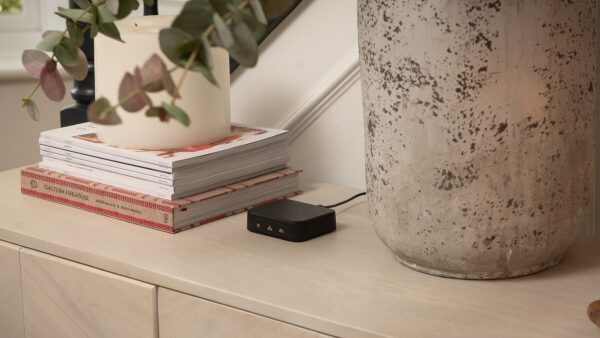 A small black ivie smart energy gateway hub with indicator lights sitting on a wooden sideboard next to books, a candle and a large vase