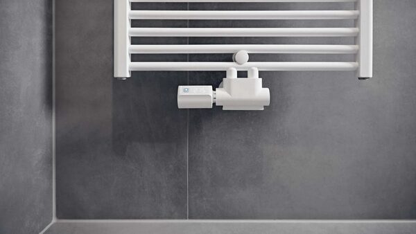 A Homematic IP radiator thermostat fitted to the valve of a white towel radiator in a grey tiled bathroom