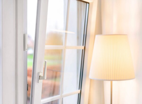 A tilted-open white uPVC window with a small white door/window contact sensor on the frame, next to a lit floor lamp