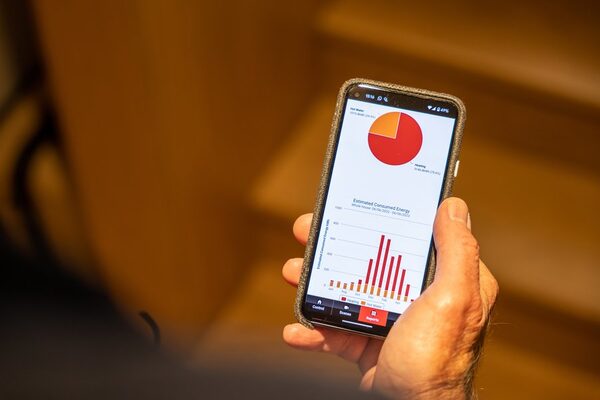 A hand holding a smartphone displaying an energy monitoring app with a pie chart and bar chart of estimated consumed energy