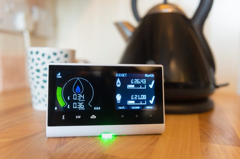 A smart meter in-home display showing energy use and cost sitting on a kitchen worktop beside a lit gas hob with a saucepan