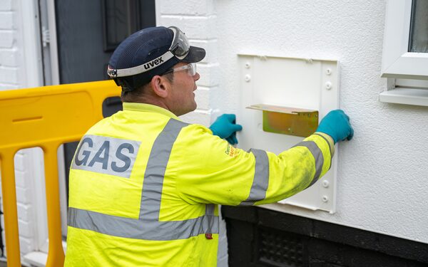 A gas engineer in a high-visibility GAS jacket and safety glasses working on a white gas meter box on an outside house wall
