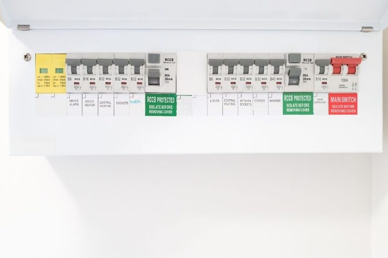 A white consumer unit (fuse box) with circuit breakers, RCD labels and a main switch mounted on a white wall
