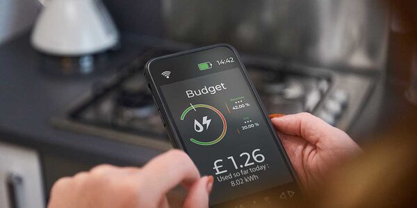 A person holding a smartphone in a kitchen showing a smart meter app with a budget gauge and £1.26 used so far today