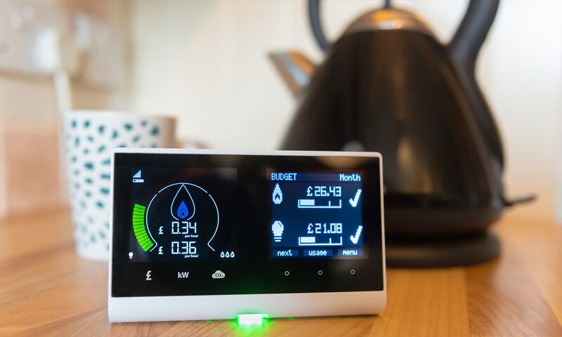 A smart energy monitor display on a kitchen worktop showing live energy use and monthly budget figures, with a kettle and mug behind