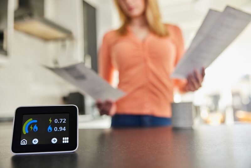 A smart meter display showing electricity and gas readings on a kitchen counter with a blurred woman holding energy bills behind