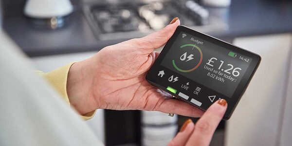 A person holding a smart meter in-home display showing energy use of £1.26 and 8.02 kWh in a kitchen