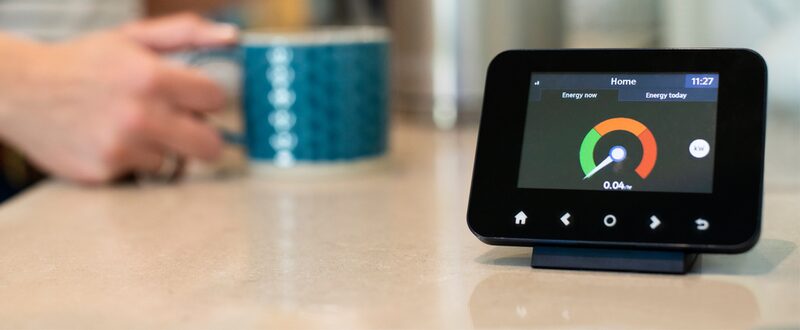 A smart meter in-home display on a kitchen worktop showing live energy usage, with a person holding a mug in the background