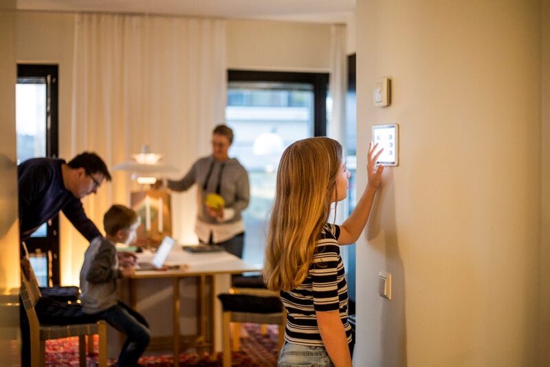 A girl touching a wall-mounted smart meter display in a home while a family sits at a table in the background