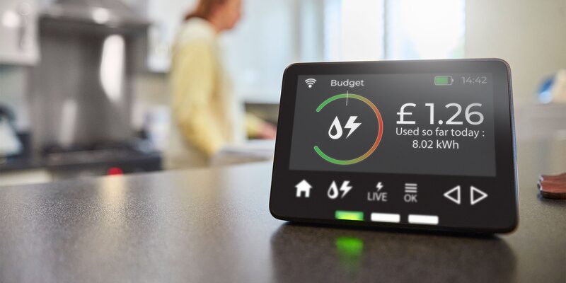 A smart energy monitor display on a kitchen worktop showing £1.26 used so far today (8.02 kWh) with a woman blurred in the background